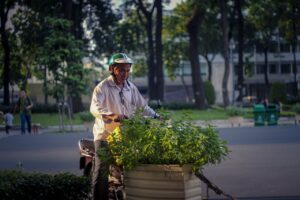 Man wearing helmet tending plants on motorcycle in urban park setting.